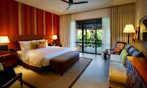 Et rummeligt soveværelse med en stor seng, to natborde med lamper, en sofa, en lænestol og en balkon med udsigt over frodig vegetation. (AI-genereret tekst)