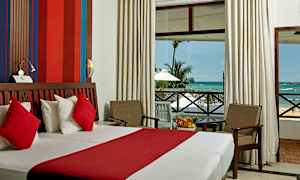 Et hotelværelse med en dobbeltseng med rødt og hvidt sengetøj, en siddegruppe med to stole og et bord, og en stor skydedør, der viser udsigten til havet og palmer. (AI-genereret tekst)