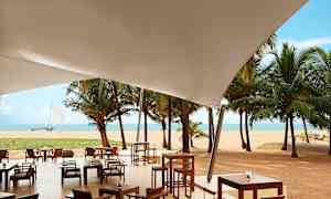 Udendørs serveringsområde under et hvidt overdækning med udsigt over sandstrand, palmer og hav. (AI-genereret tekst)