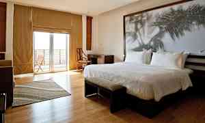 Et moderne hotelværelse med en stor seng, skrivebord, stol og en stor glasdør, der fører til en balkon. (AI-genereret tekst)