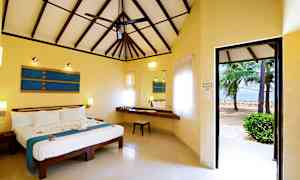 Et soveværelse med højt loft og en ventilator. Værelset har en stor seng med blå dekorative detaljer, et skrivebord med en stol samt vinduer med lyse gardiner. Døren åbner ud til en strand med palmer. (AI-genereret tekst)