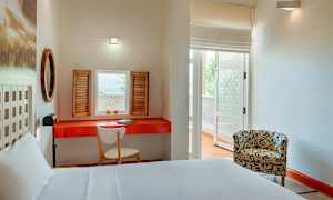 Et lyst hotelværelse med en hvid seng, et rødt bord med en skrivebordsstol og skodder ved vinduet. En mønstret lænestol står på den anden side af rummet. (AI-genereret tekst)