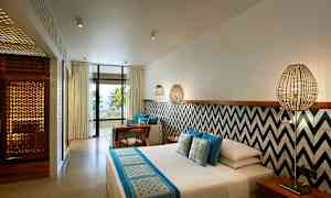 Et hotelværelse med moderne indretning, seng med blå og hvide tekstiler, væg med geometriske mønstre, siddeområde og en balkon med udsigt til palmer og havet. (AI-genereret tekst)