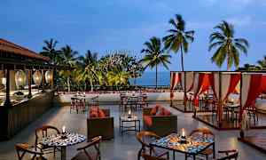 En udendørsrestaurant med borde, stole og sofaer, omgivet af palmer med havet synligt i baggrunden. (AI-genereret tekst)