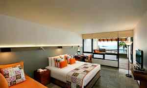 Hotelværelse med moderne indretning, indeholdende en dobbeltseng med farverige puder og sengetæppe. Et skrivebord, en sofa og et fjernsyn kompletterer rummet. Der er en balkon med udsigt over havet. (AI-genereret tekst)