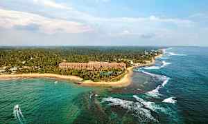 Luftfoto af en tropisk kystlinje med et hotel tæt på stranden, omgivet af palmer og havet. (AI-genereret tekst)