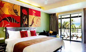 Hotelværelse med en stor seng, farverige puder, en siddegruppe og en balkon med udsigt over en sandstrand omgivet af palmer. (AI-genereret tekst)