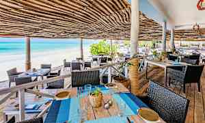 Udendørs serveringsområde med tag af flettet bast ved en sandstrand og blåt hav. (AI-genereret tekst)