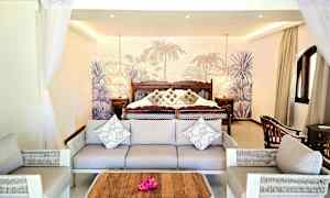 Et hotelværelse med sofa og lænestole foran en stor seng med pyntepuder og tropisk baggrund. (AI-genereret tekst)