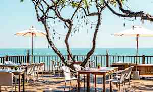 En udendørsrestaurant med træborde og stole på en terrasse med udsigt over havet. Et stort træ med lyskæder står i centrum. (AI-genereret tekst)