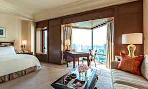 Et elegant hotelværelse med en stor seng, en sofa, et skrivebord og store vinduer med udsigt over byen. (AI-genereret tekst)