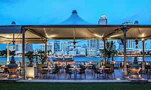 Udendørs servering med borde og stole, beliggende ved en flod, med en by i baggrunden. (AI-genereret tekst)