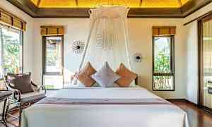 Et soveværelse med en stor seng dekoreret med puder og en hvid baldakin. Værelset har vinduer med udsigt til grønne omgivelser. (AI-genereret tekst)