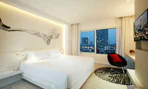 Et moderne hotelværelse med en stor seng, et vindue med udsigt over bybygninger, en stol og et tv. (AI-genereret tekst)