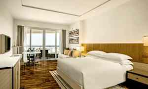 Et moderne hotelværelse med en stor seng, sofa og spisebord i et lyst lokale. Værelset har en balkon med udsigt og et fjernsyn på væggen. (AI-genereret tekst)