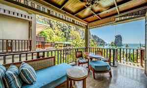 En terrasse med siddeområde under tag, med udsigt over en frodig tropisk natur og havet i baggrunden. (AI-genereret tekst)