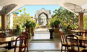 Udendørs servering med træstole og borde, omgivet af grønne områder og dekorative planter. En arkitektonisk struktur ses i baggrunden. (AI-genereret tekst)