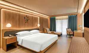 Et moderne hotelværelse med en stor seng, natbord, en lænestol, et skrivebord og et stort vindue med gardiner. (AI-genereret tekst)