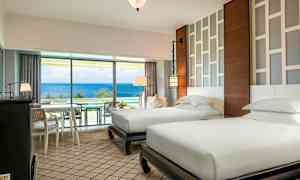 Et hotelværelse med to separate enkeltsenge, moderne møbler og et lyst design. Et stort vindue med udsigt over havet. (AI-genereret tekst)