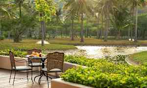 Udendørs serveringsområde med bord og stole på en træterrasse, omgivet af grønt og udsigt over en dam. (AI-genereret tekst)