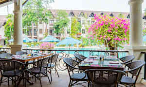 Udendørs servering med borde og stole ved en pool. I baggrunden ses bygninger, parasoller og grønne områder. (AI-genereret tekst)