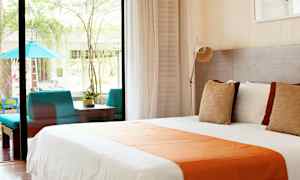 Billede af et lyst hotelværelse med en stor seng dækket med hvidt og orange sengetøj, store vinduer, der åbner ud til en terrasse med siddeområde og planter. (AI-genereret tekst)