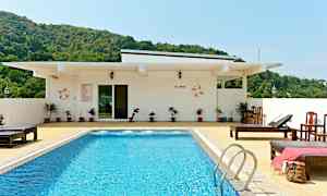 En pool med blåt vand omgivet af solsenge og en hvid bygning i baggrunden. Grønnere bakker er synlige bag bygningen. (AI-genereret tekst)