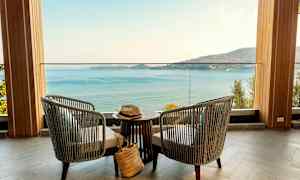 To stole og et bord står på en terrasse med udsigt over havet og en bjergkyst. (AI-genereret tekst)