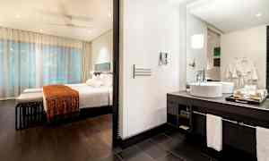 Et hotelværelse med soveområde og badeværelse. Soveområdet har en seng med hvidt sengetøj og orange tæppe. Badeværelset har en håndvask med spejl. (AI-genereret tekst)