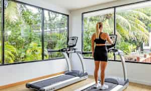 En person ser på skærmen på et løbebånd i et motionscenter med store vinduer og udsigt til grønne områder. (AI-genereret tekst)