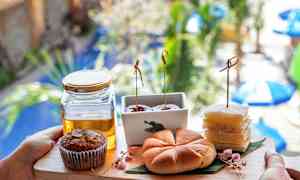 En bakke med forskellige bagværk og en glasbeholder med honning, holdt foran en uklar baggrund med planter og parasoller. (AI-genereret tekst)