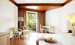 Et hotelværelse med hvidt skrivebord, seng og siddepladser samt altan med udsigt over træer. (AI-genereret tekst)