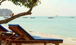 To solsenge med blå hynder på en sandstrand ved havet, med både i baggrunden. (AI-genereret tekst)