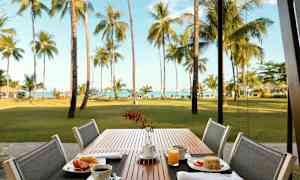 Udendørs spisested med en morgenmad ved et bord. Baggrunden viser en græsplæne, palmer og hav. (AI-genereret tekst)