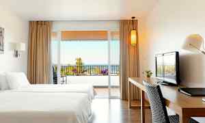 Et hotelværelse med to senge, skrivebord og udsigt til en altan med hav i baggrunden. (AI-genereret tekst)