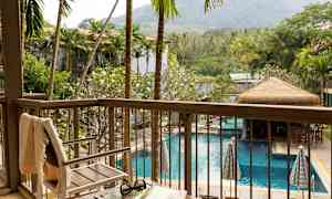 Udsigt fra en altan over en pool omgivet af palmer og grønt, med en stol og et bord i forgrunden. (AI-genereret tekst)