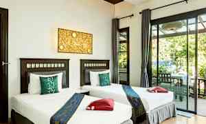 To enkeltsenge med hvidt sengetøj og blå dekorative sengetæpper står side om side i et lyst rum med glasdøre, der åbner ud til en terrasse med udsigt til grønne omgivelser. (AI-genereret tekst)