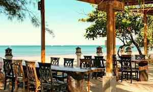 En udendørsrestaurant med træborde og stole, med udsigt over en sandstrand og havet. (AI-genereret tekst)