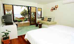 Et lyst hotelværelse med seng, sofa og en stor glasdør, der fører ud til en terrasse med hængekøje og udsigt over havet. (AI-genereret tekst)