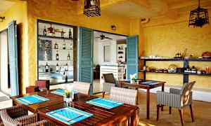 Et rum med gule vægge og turkise døre, indrettet med træborde og fletstole. Hylder med flasker og serveringsartikler er synlige. (AI-genereret tekst)