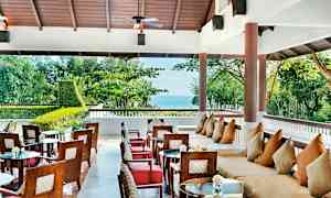 En åben siddegruppe med borde og stole samt sofaer med puder, placeret nær grønne træer med udsigt over havet. (AI-genereret tekst)