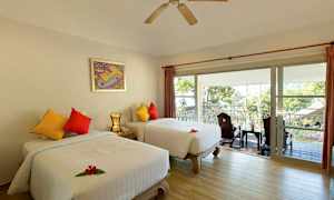 Et hotelværelse med to separate senge, farverige puder og en balkon med udsigt over træer og et tag udenfor. (AI-genereret tekst)