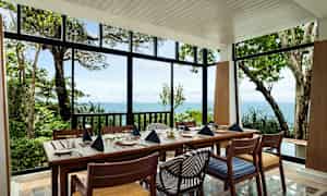 Et dækket spisebord med stole i en lys pavillon med udsigt over havet og frodig natur. (AI-genereret tekst)