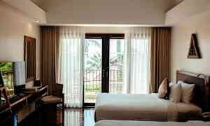 Et hotelværelse med to senge, et skrivebord med stol, og store vinduer med udsigt til en altan og træer udenfor. (AI-genereret tekst)