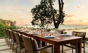 Et dækket bord med stole står udendørs ved solnedgang med hav og træer i baggrunden. (AI-genereret tekst)