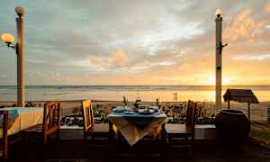 Udsigt over en strand ved solnedgang, med et dækket bord og stole i forgrunden. (AI-genereret tekst)