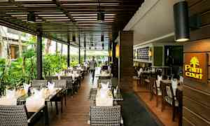 En restaurant med udendørs servering, indrettet med kurvestole og dækkede borde med duge og glas. (AI-genereret tekst)