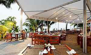 En udendørsservering med træborde og stole under et hvidt stofloft, omgivet af tropiske palmer. (AI-genereret tekst)