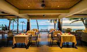 En restaurant ved havet med hvide duge på bordene og udsigt over palmer og stranden. (AI-genereret tekst)