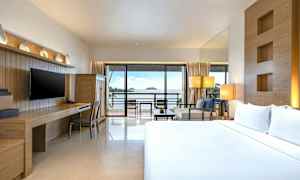 Et hotelværelse med en dobbeltseng, skrivebord og tv samt udsigt over havet gennem en stor glasdør, der fører til en altan. (AI-genereret tekst)